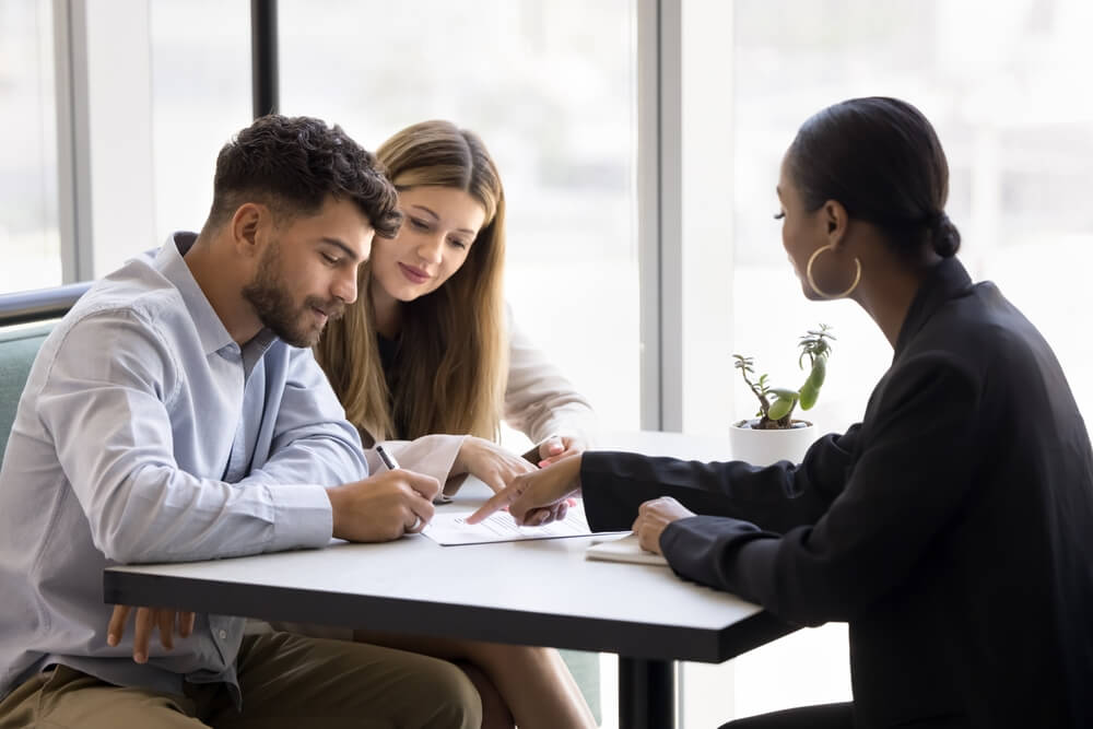 Confident young African realtor agent pointing at paper agreement for couple of customers, clients signing, giving consultation, legal help with mortgage, real estate property buying