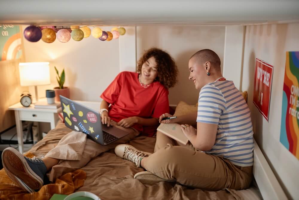 Portrait of two cheerful young students using laptop together sitting on bunk bed in college dorm