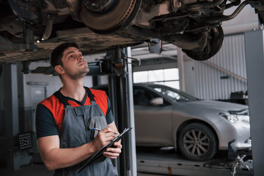 ar mechanic taking a look at car undercarriage and transmission while writing notes in a pad