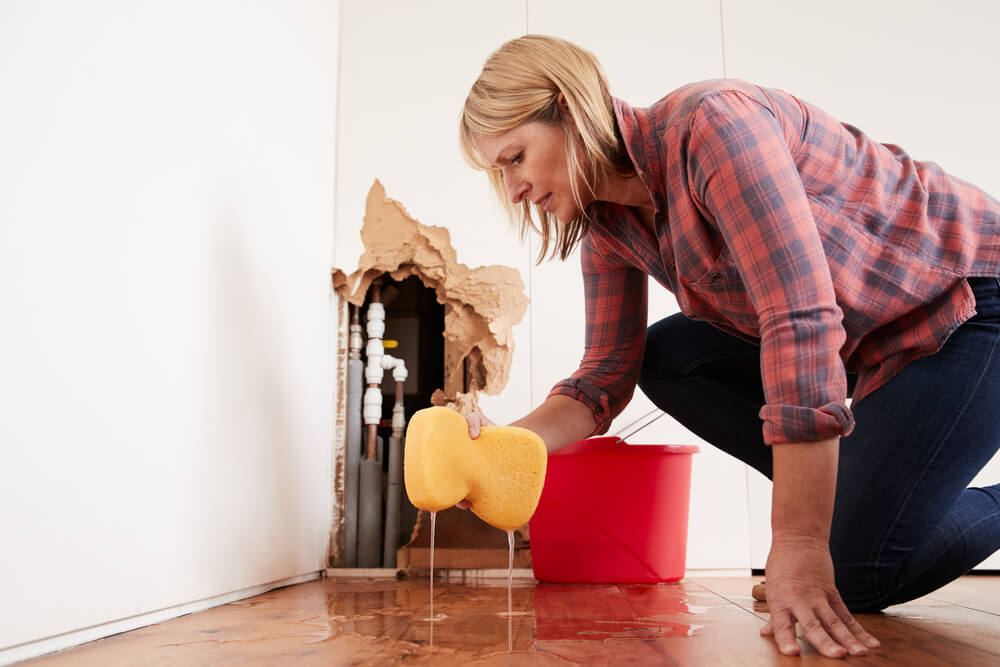 A woman in a red plaid shirt squatting beside a burst pipe that has created a hole in the wall. She is swabbing with a sponge and bucket.