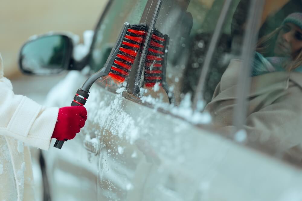 Driver Uses a Snow Broom to De-ice the Car. Woman getting her car ready for commute in a winter morning