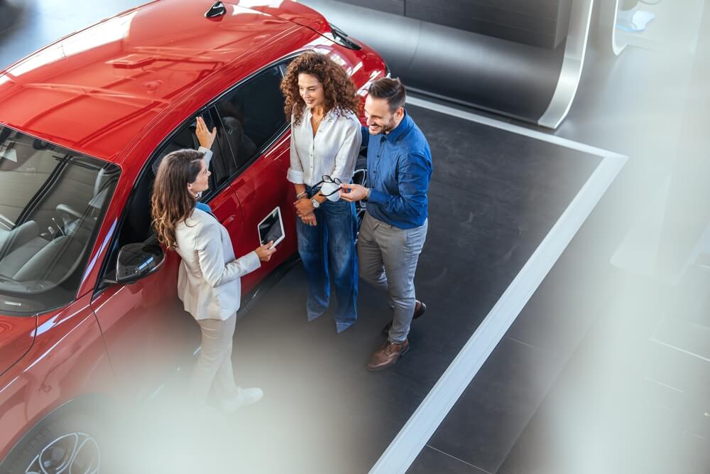 A young professional couple at a dealership being shown a red car by a salesperson