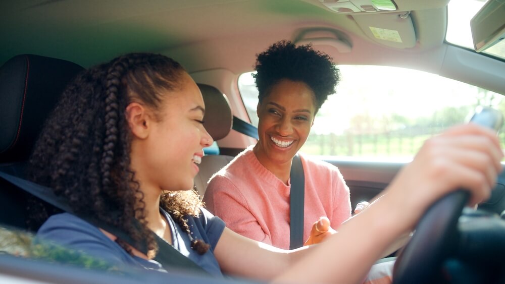 A teenage driver behind the wheel smiling next to her mother in the passenger seat