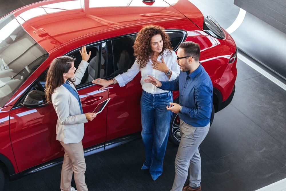 A friendly saleswoman discusses car options with a young couple beside a shiny red vehicle at a dealership