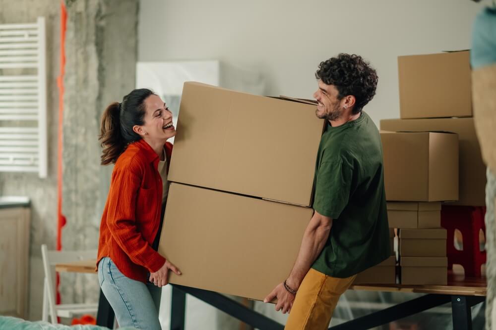 Happy couple carrying stacked cardboard boxes while moving into new apartment