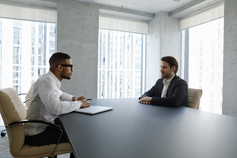 Side view concentrated young professional African American professional hr manager or employer holding paper CV in hands, listening qualified male job seeker, sitting together at table in office.