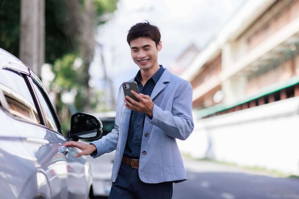 Young asian man smiling, using a smartphone app while opening a car door