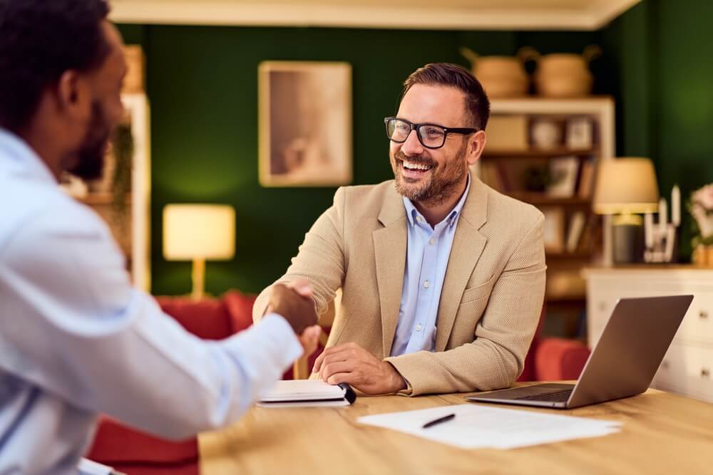 Two professionals seal an agreement with a handshake in a cozy office, with laptop and documents nearby