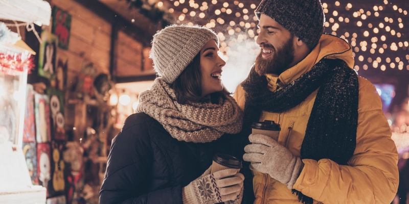 A man and woman in winter attire hold hot drinks and walk through a holiday market