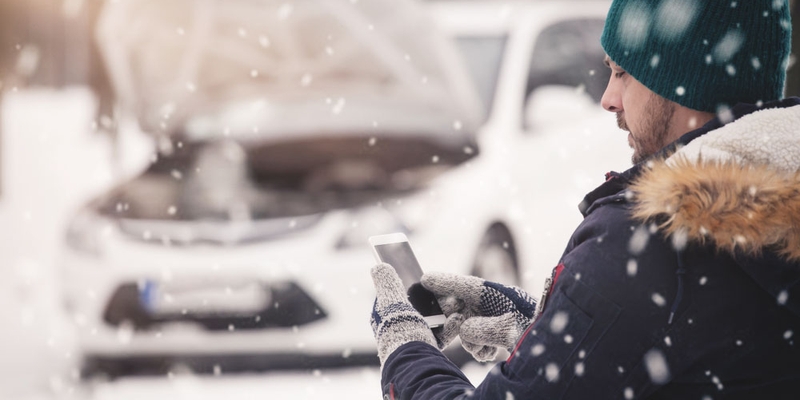 A man in winter clothing uses a cell phone and calls for assistance for his broken down vehicle