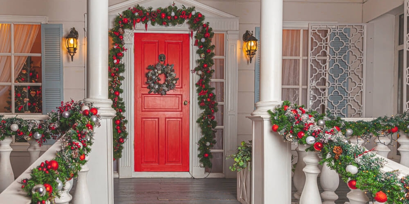 House entrance with red door decorated for holidays. Red and green wreath garland of fir tree branches and lights on railing. Christmas eve at home. (1).jpg