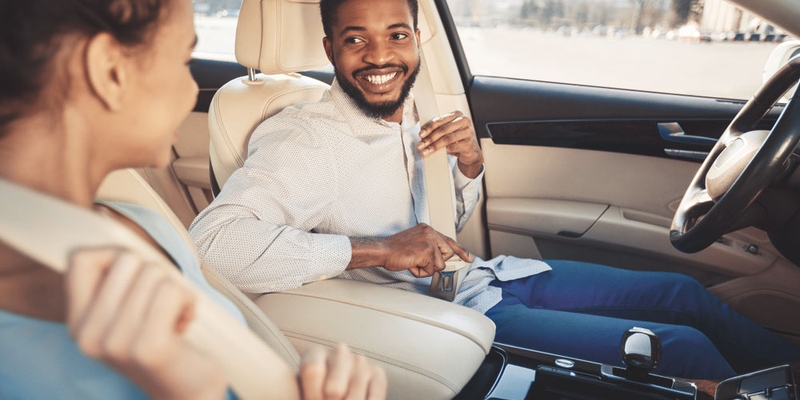 young man buckles seat belt with friend in car smiling