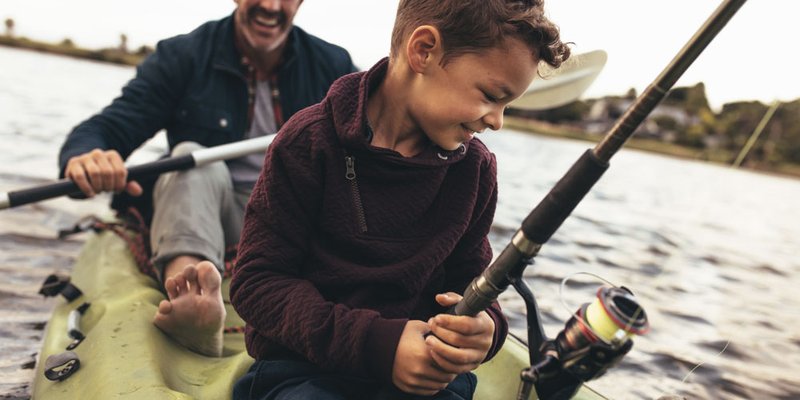 A dad paddles a little boat while the son sits up front and tries to fish