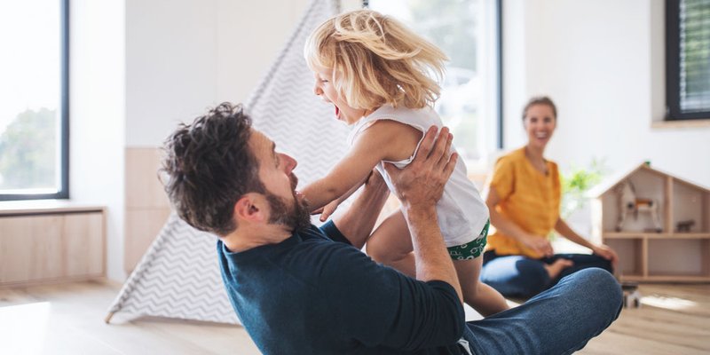 A dad plays with his young daughter at home in their living room while mama laughs happily in the background