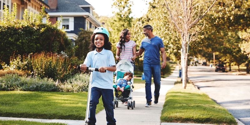 A family walks down the neighbourhood street while their child gets ahead on the scooter