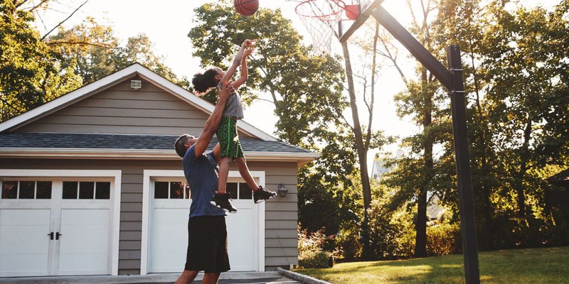 A dad picks up his daughter and holds her up high so she can score a basket at their driveway basketball hoop