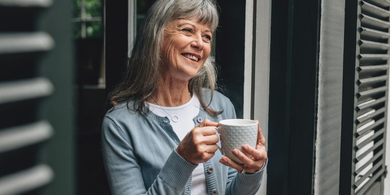 A senior woman enjoys a cup of coffee while standing outside on her porch