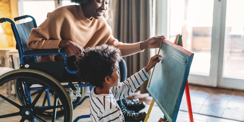 Woman in wheelchair helping child write on chalkboard