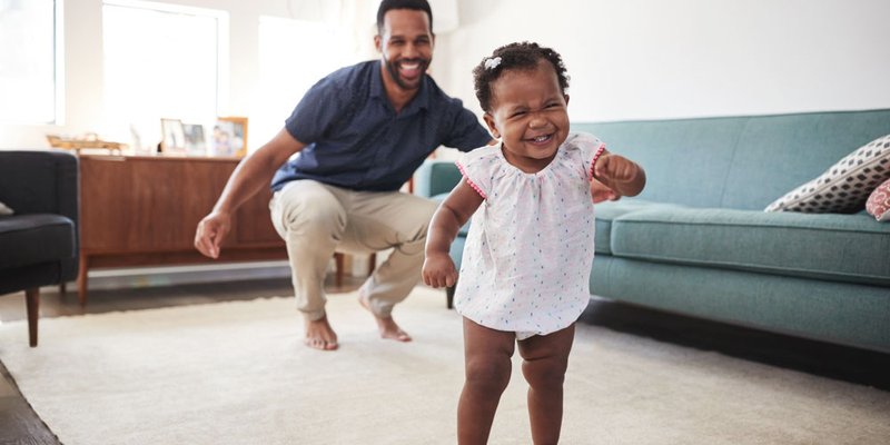 Proud papa watches baby girl learn to walk in living room