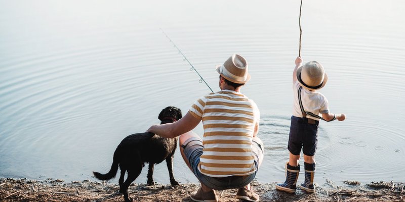 Dad and son in matching hats fish together with their dog at a nice still pond