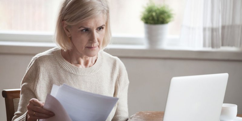 An older woman looking through documents as she works on her laptop