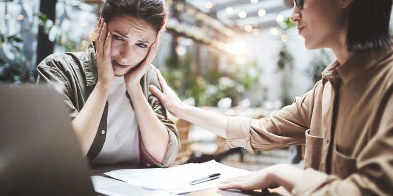 A woman comforts another woman at a table with a laptop and documents