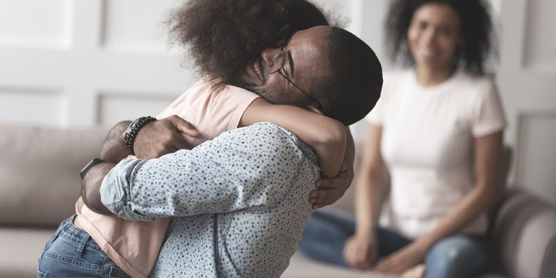 A father hugs his daughter in the foreground and the mother is blurred out in the background