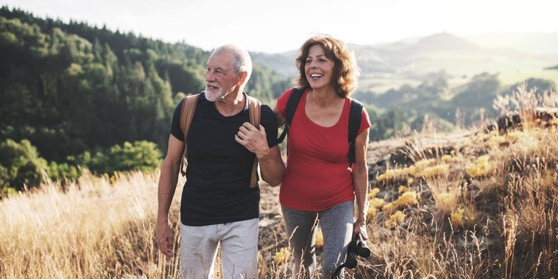 An older man and woman carry backpacks and walk outdoors