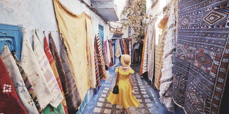 A woman in a flowing yellow dress walks down a marketplace with rugs hanging