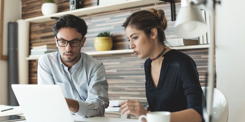 A man and a woman work together at a desk with a laptop and notebook