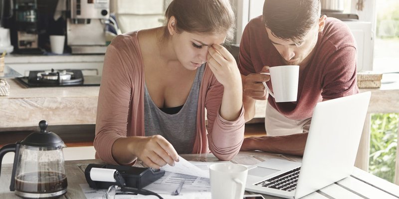A man and woman sit at a table covered in papers and look worried