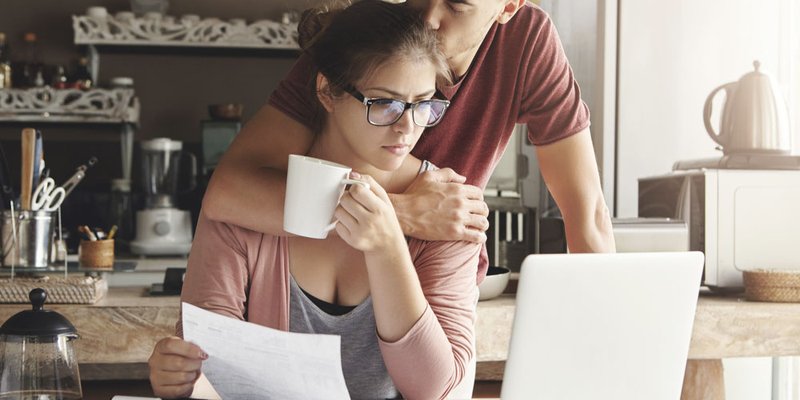 A man kisses a woman's forehead as she looks over her bills at the kitchen table