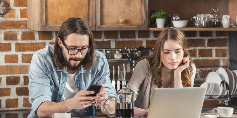 A man sits using his smartphone next to a woman using her laptop