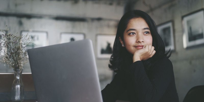Young woman sits in front of a computer and softly smiles while looking off into the distance.
