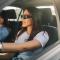 Portrait of young trendy attractive female driver driving a car with hands on steering wheel and smiling while her friend is sitting next to her and enjoying a ride. Two friends in car on a road trip.