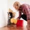 A woman in a red plaid shirt squatting beside a burst pipe that has created a hole in the wall. She is swabbing with a sponge and bucket.