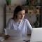 Focused busy young office employee working at laptop, reviewing online reports, checking financial documents, holding paper contracts, insurance agreement. Business woman doing paperwork job