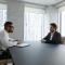 Side view concentrated young professional African American professional hr manager or employer holding paper CV in hands, listening qualified male job seeker, sitting together at table in office.