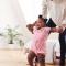 A mama helping her baby girl learn to walk towards her dada at home in their living room