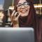 A young woman happily chats on speaker phone while working on her laptop in a window seat somewhere downtown