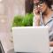 A professional woman in trendy glasses sits outside at a table while working on her laptop and talking on the phone