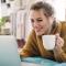 Woman lays on bed casually browsing on her laptop and drinking a coffee
