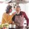 An older woman lets a man taste sauce off a wooden spoon in the kitchen